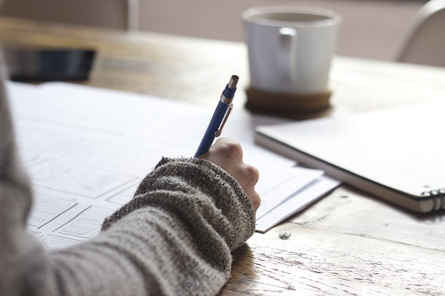 Person writing on paper with a pen, cup of coffee in the background, illustrating the process of applying for bank statement loans for self-employed individuals.