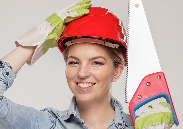 Woman wearing a red hard hat and work gloves, holding a handsaw, smiling, representing renovation and construction themes relevant to FHA 203K loans.